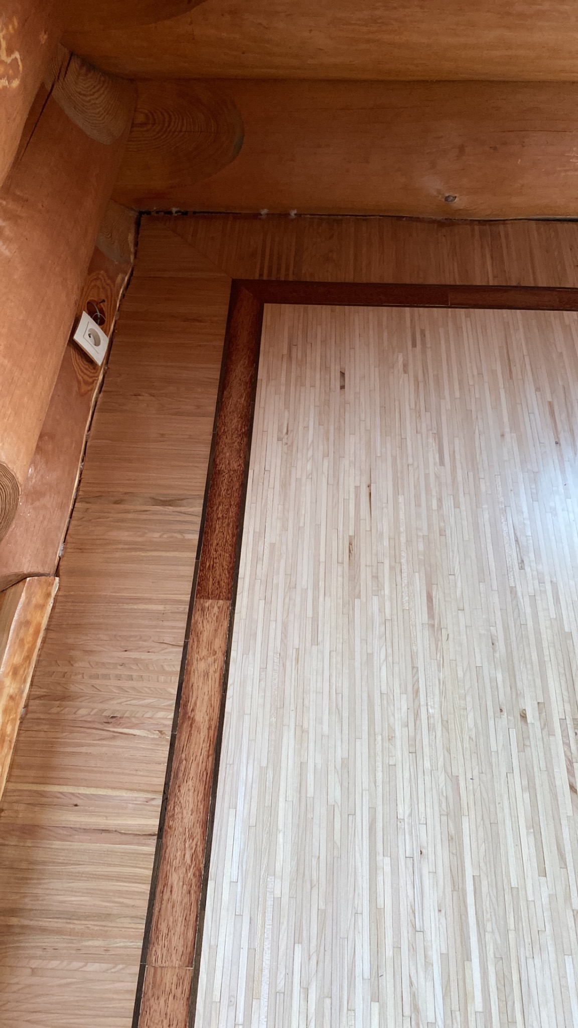 Fragment drewnianego wnętrza z wykończoną podłogą z jasnych desek, obramowaną ciemniejszą listwą przypodłogową, widoczny fragment ściany i sufitu z drewnianych bali, gniazdko elektryczne na ścianie.