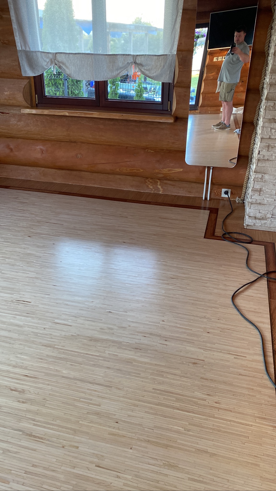 Jasny, drewniany parkiet w rustykalnym wnętrzu z bali, widoczne okno z zasłonami i odbicie osoby w lustrze.