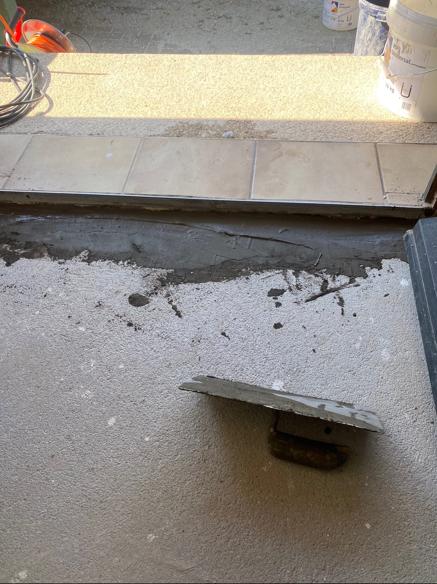 Przygotowanie podłoża pod płytki: warstwa zaprawy wyrównującej, widoczna paca, fragment ułożonych płytek i wiaderka z materiałami budowlanymi w tle.