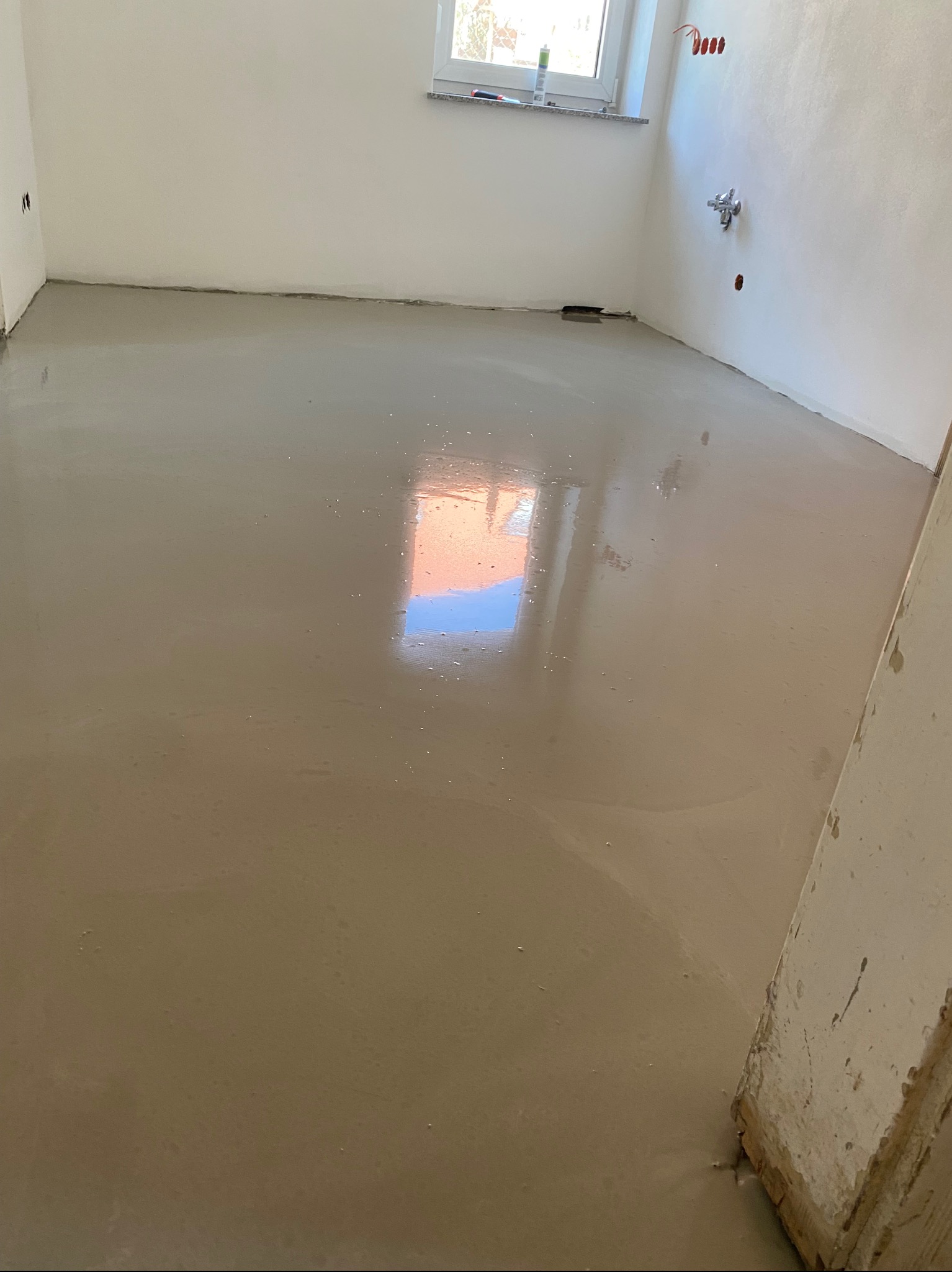 Świeżo wylana, samopoziomująca wylewka podłogowa w jasnym, pustym pokoju z oknem i instalacją wodną na ścianie.