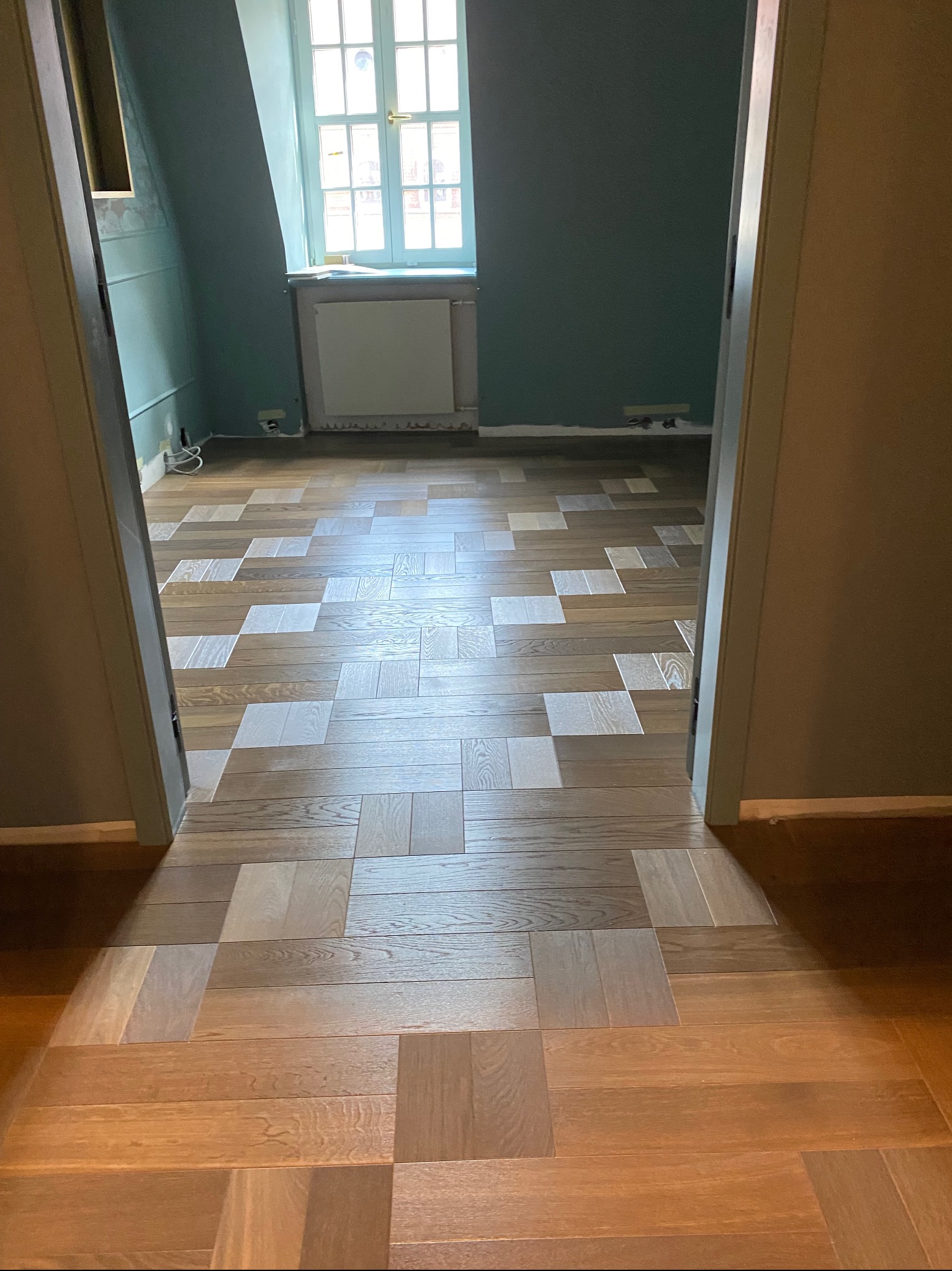 Ukończona podłoga z ułożonych parkietów w geometrycznym wzorze, łącząca ciemne i jasne drewno, widziana z perspektywy wejścia do pokoju z zielonymi ścianami.
