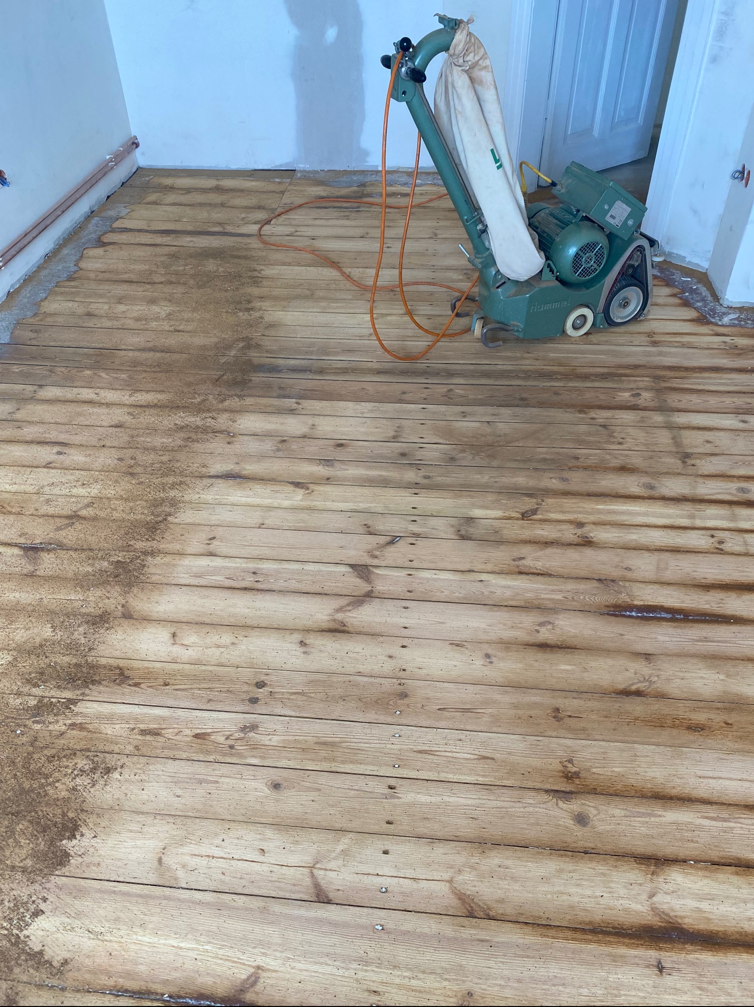 Zielona szlifierka do parkietu Hummel stoi na częściowo wyszlifowanej drewnianej podłodze; widoczne ślady pyłu i różnica w odcieniu drewna przed i po szlifowaniu.