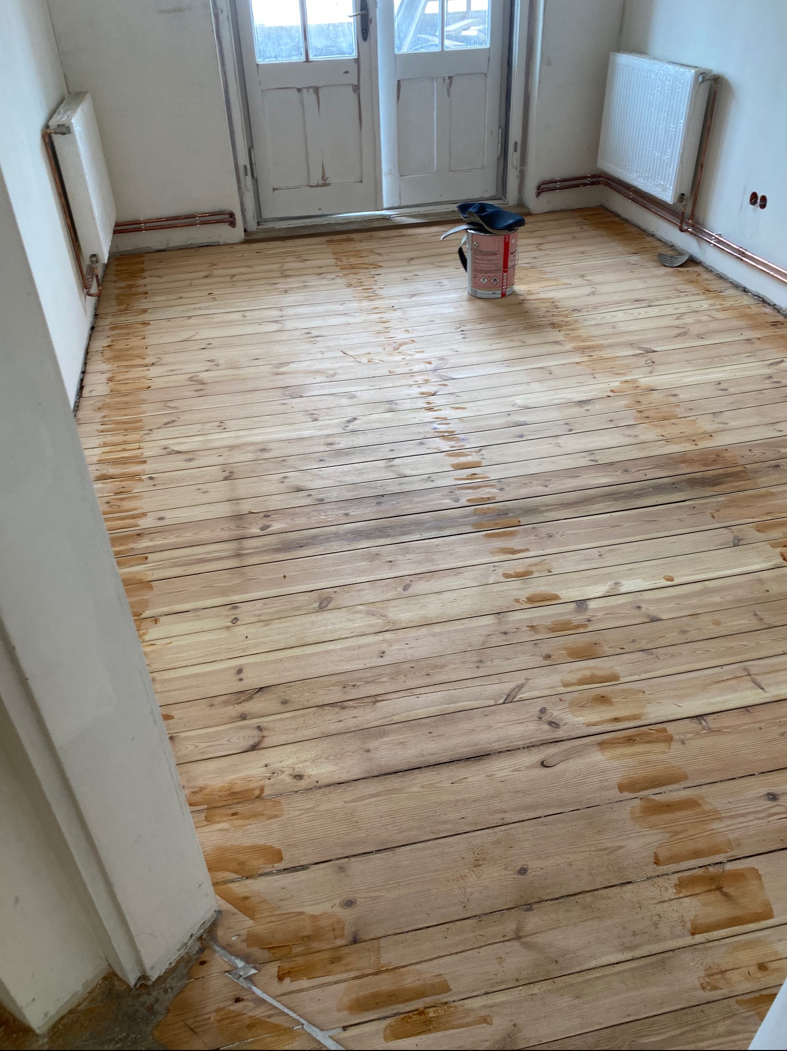 Drewniana podłoga po częściowym nałożeniu bejcy, widoczne ślady po pędzlu, puszka z bejcą i zabezpieczony grzejnik w tle.
