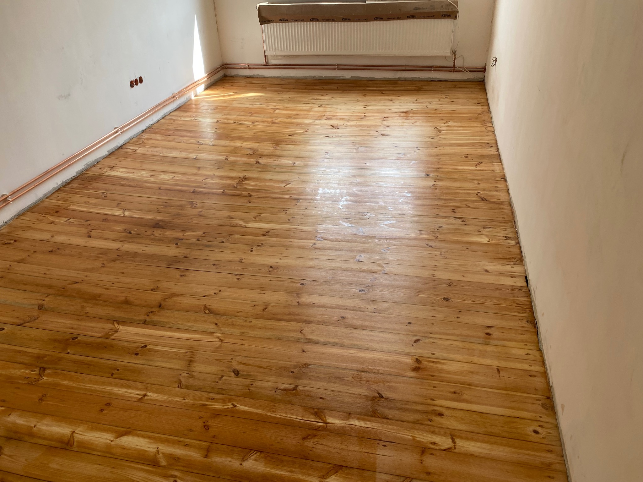 Świeżo polakierowana drewniana podłoga z widocznymi miedzianymi rurami grzewczymi przy ścianie i grzejnikiem w tle, w trakcie remontu pomieszczenia.