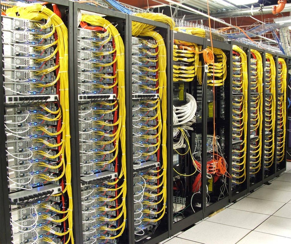 Rząd serwerów w serwerowni z żółtymi kablami, widoczne złącza i kable w różnych kolorach. Sprzęt IT w pomieszczeniu z podwieszanym sufitem.
