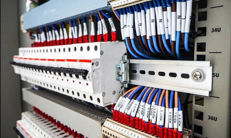 Rozdzielnica elektryczna z bezpiecznikami i okablowaniem oznaczonym etykietami, widok z bliska na instalację w szarej obudowie.