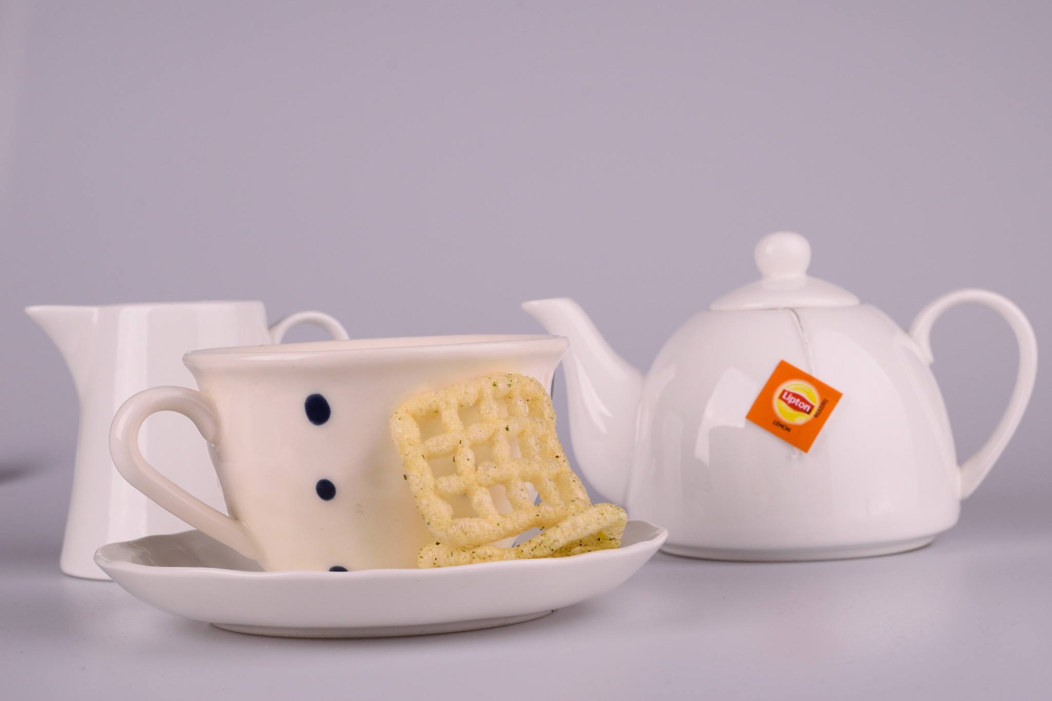 Biała filiżanka w niebieskie kropki na spodku, z waflem w kształcie krzesła obok, dzbanek na mleko i czajnik z pomarańczową etykietą Lipton w tle, na szarym tle.