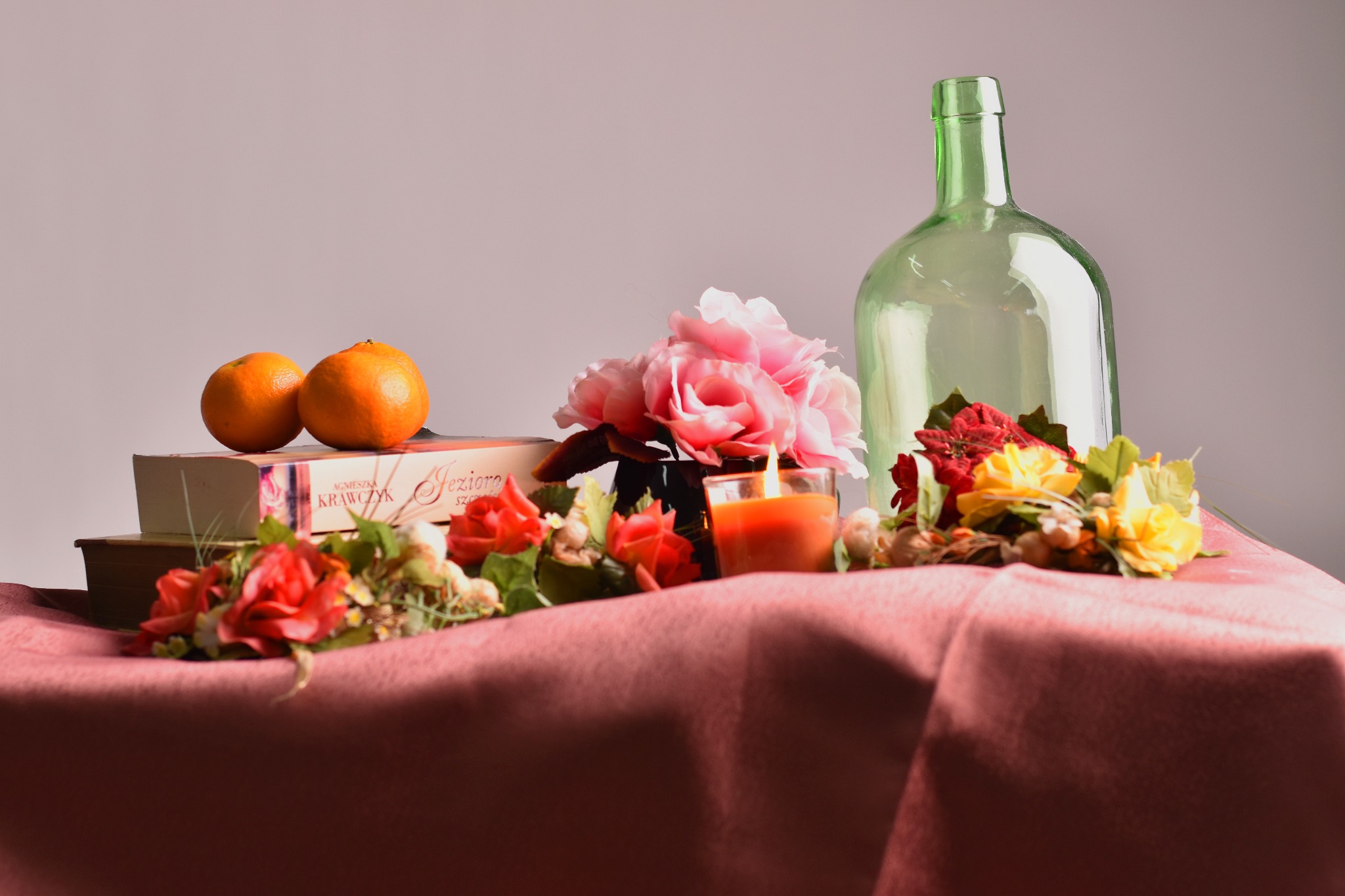Martwa natura: dwie pomarańcze na książkach, świeca w szklanym naczyniu, bukiety kwiatów i zielona butelka na różowym obrusie.