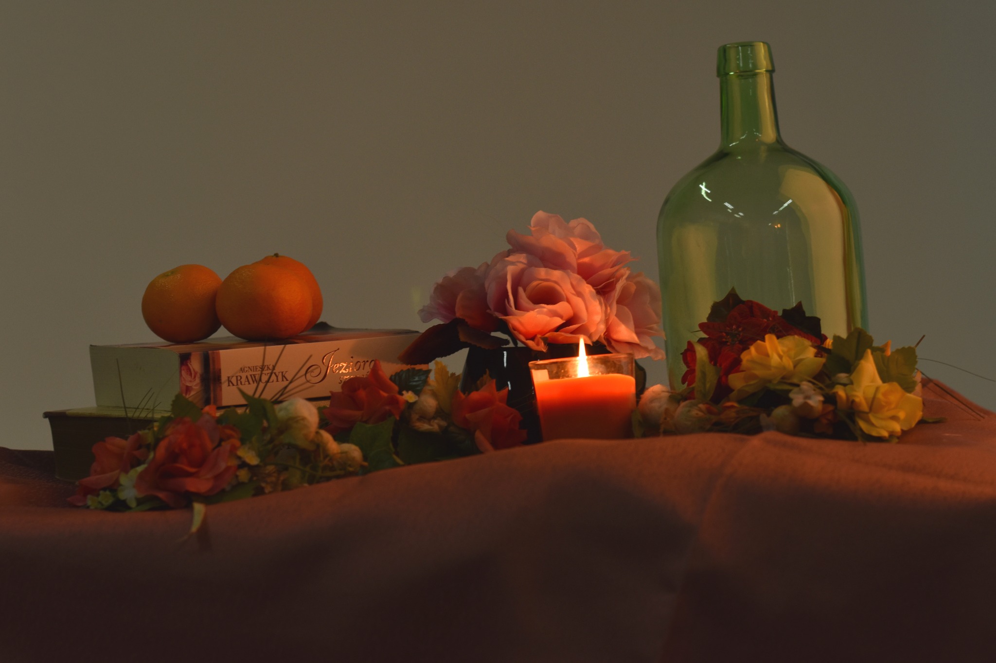 Martwa natura: dwie pomarańcze na książkach, bukiet różowych i czerwonych kwiatów, paląca się pomarańczowa świeca w szklanym naczyniu i zielona butelka na tle szarego tła.