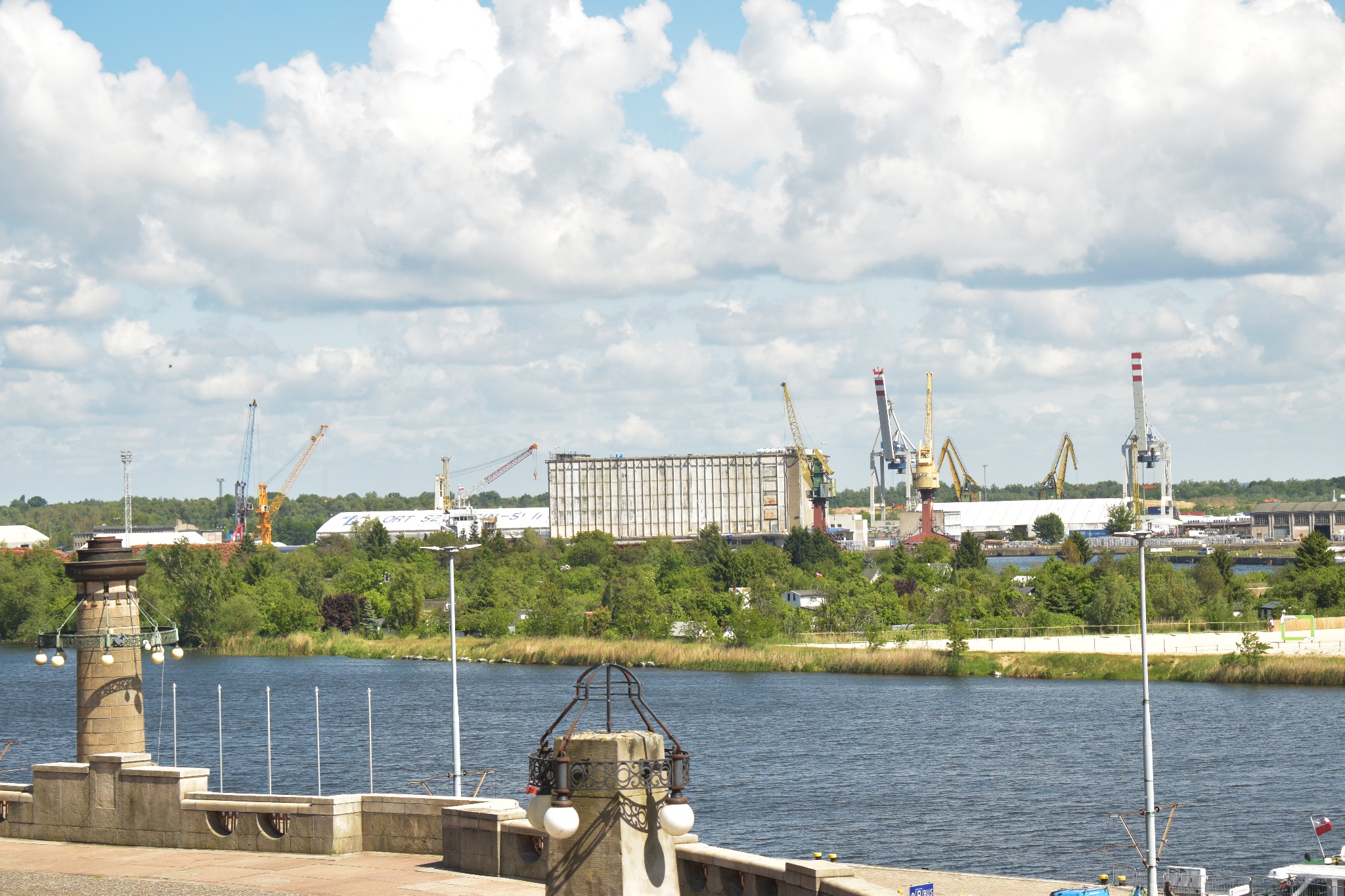 Widok na port z rzeką, dźwigami i magazynem na drugim brzegu, uchwycony z kamiennej balustrady z ozdobną latarnią.