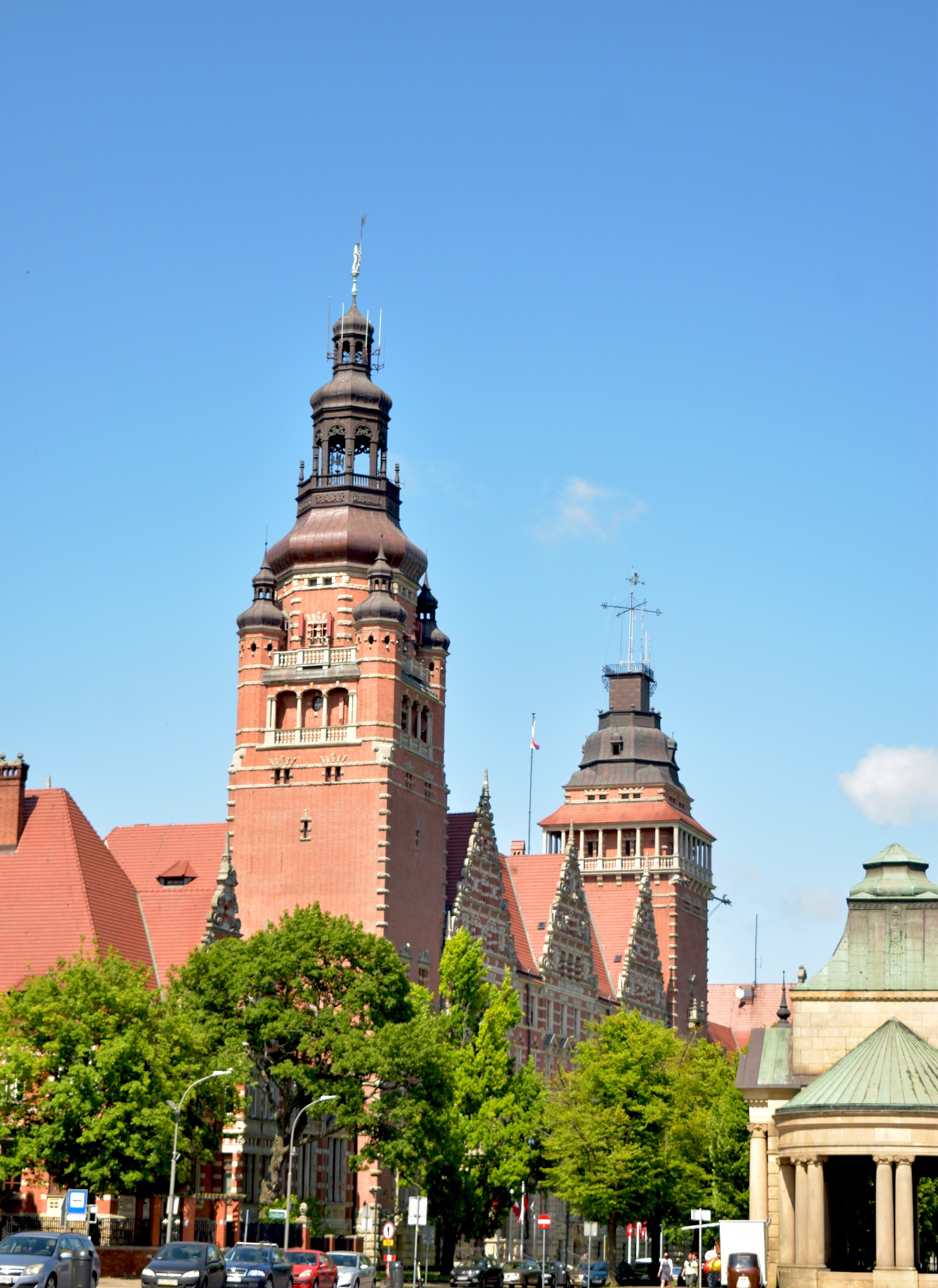 Widok na dwie wieże Muzeum Narodowego w Szczecinie z czerwonej cegły, z zielonymi drzewami w tle i fragmentem ulicy z samochodami w dolnej części kadru.