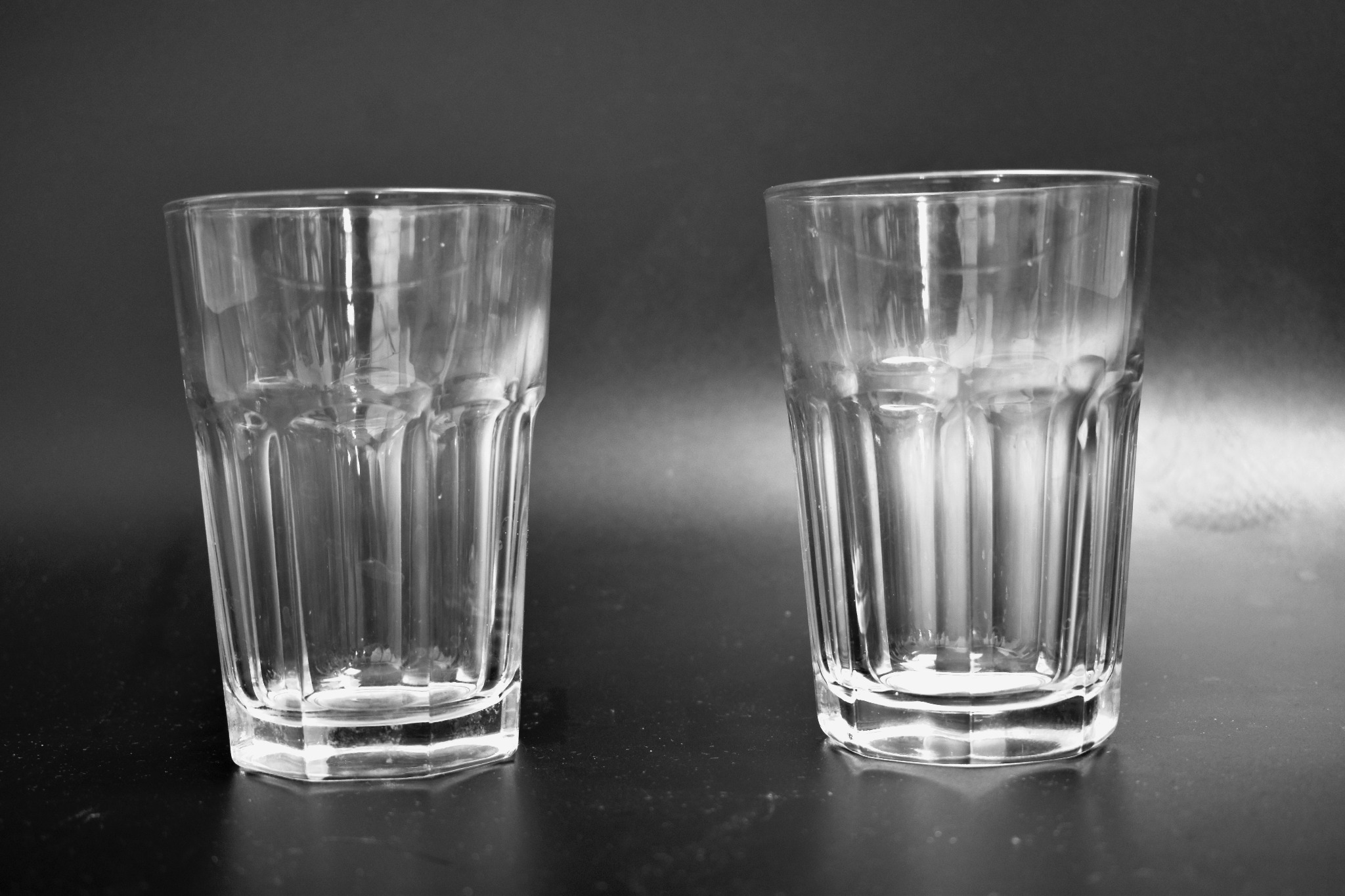 Dwa puste, szklane kubki o fasetowanej powierzchni, ustawione obok siebie na ciemnym, błyszczącym blacie, sfotografowane w czerni i bieli.