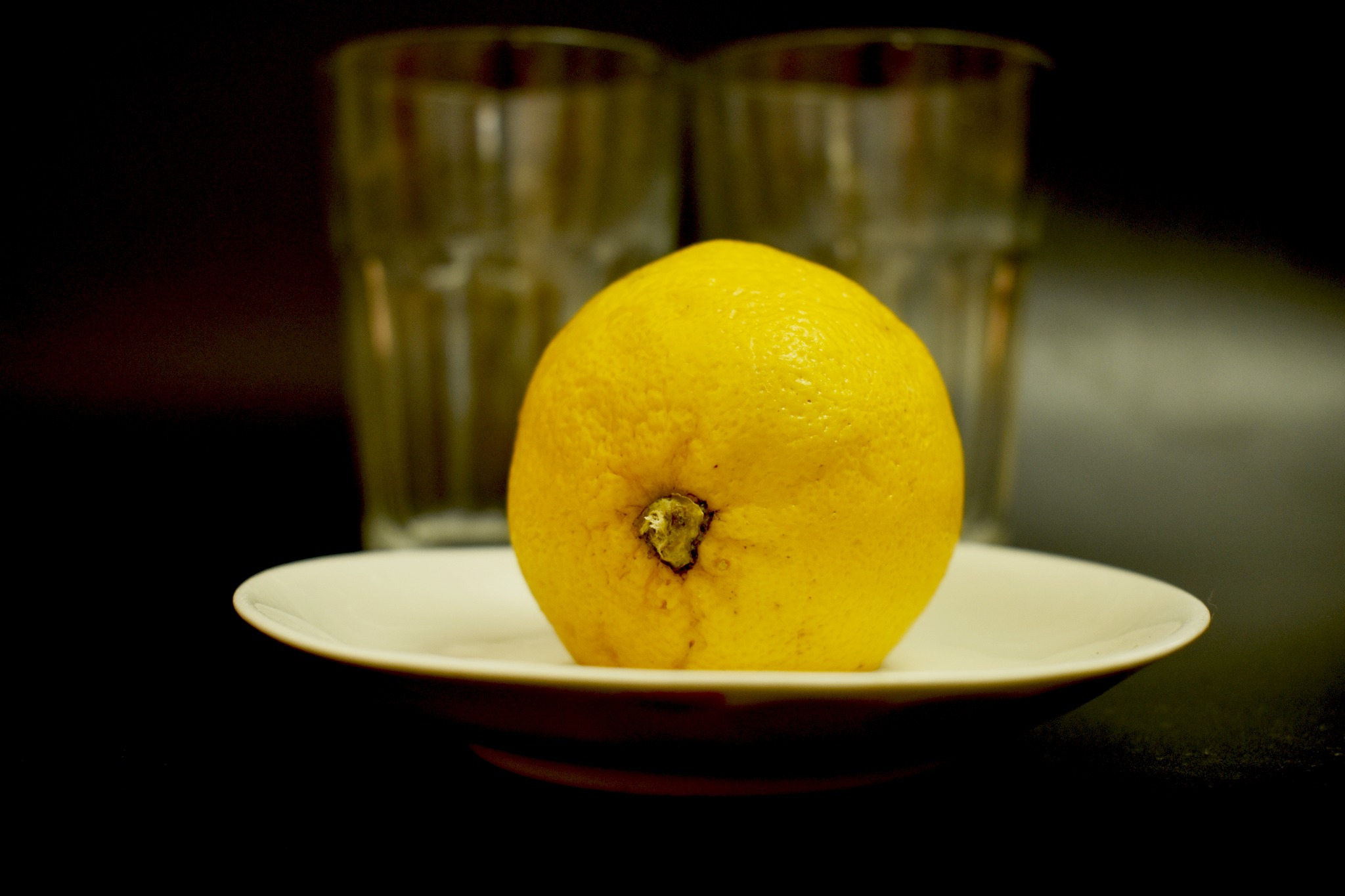 Żółta cytryna na białym talerzu, w tle dwa rozmyte szklanki, ciemne tło.