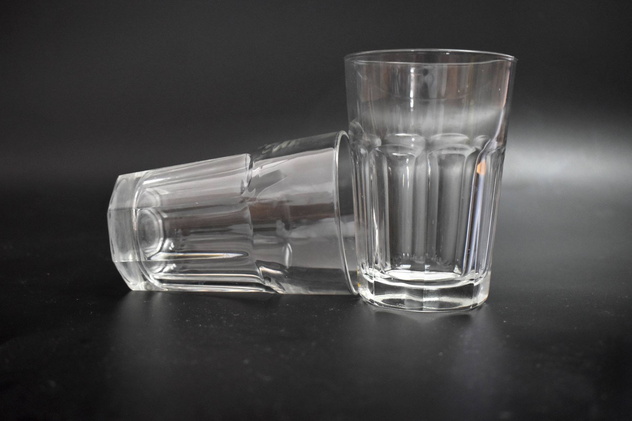 Dwa szklane kubki o prążkowanej fakturze, jeden stojący pionowo, drugi leżący na boku, oba częściowo wypełnione wodą, na ciemnym tle.