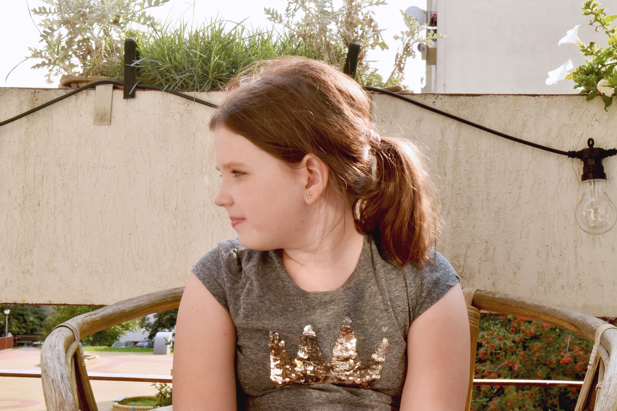 Dziewczynka z rudymi włosami spiętymi w kucyk, siedząca na drewnianym krześle na balkonie z widokiem na park, ubrana w szarą koszulkę z brokatową koroną, patrzy w lewo, w tle widać dekoracyjne...