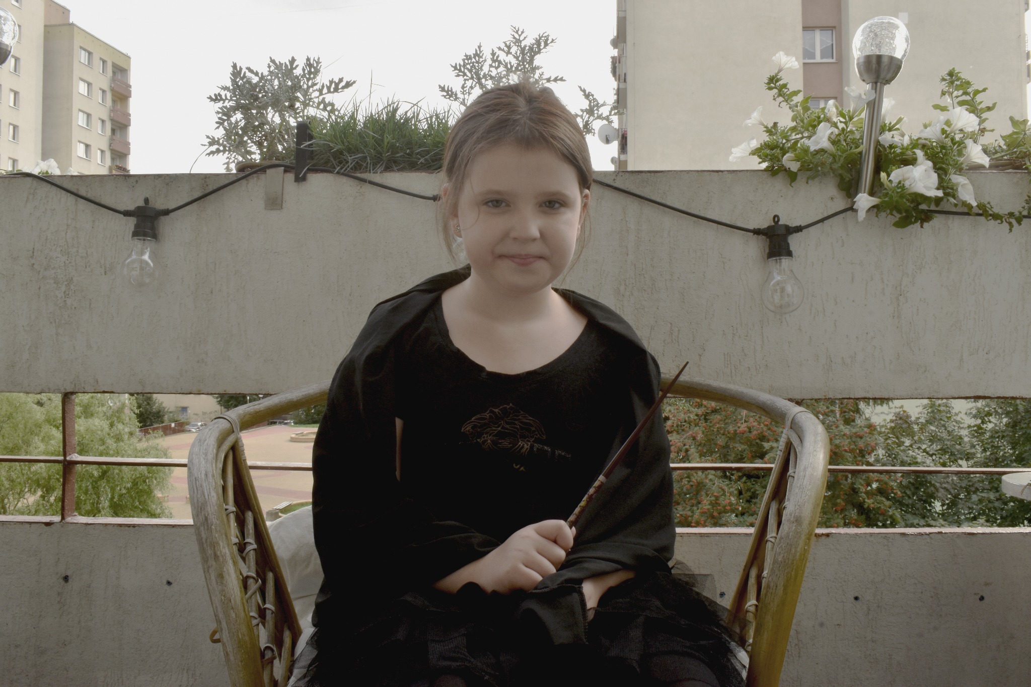 Dziewczynka w czarnej sukience siedzi na balkonie, trzymając różdżkę, w tle budynki mieszkalne i dekoracyjne oświetlenie.