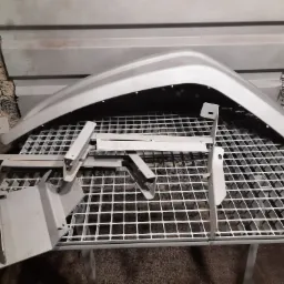 Metalowe elementy, w tym błotnik, po obróbce na metalowej kratownicy, widoczne ślady niedoskonałości na ścianie w tle.