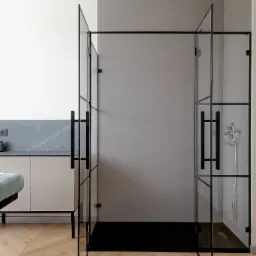 Nowoczesna kabina prysznicowa z czarnymi profilami szklanymi, widoczne uchwyty i deszczownica, na podłodze parkiet w jodełkę.