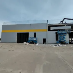 Montaż ścian hali przemysłowej z płyt warstwowych, widoczne podnośniki nożycowe i fragment dźwigu, zabezpieczenie siatką na krawędzi dachu.