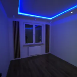 Pusty pokój z drewnianą podłogą, białymi ścianami i oknem, oświetlony niebieskim światłem LED z sufitu, z dekoracyjnymi panelami drewnianymi po bokach okna.