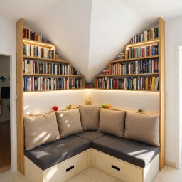 Najmart Sp. z o.o. - Kącik czytelniczy na poddaszu w Częstochowie: drewniane regały na książki z oświetleniem LED, siedzisko z szufladami i poduszkami, dopasowane do skosu dachu.