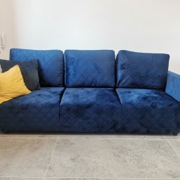 Najmart Sp. z o.o. - Granatowa sofa z pikowaniem w romby, z żółtą i granatową poduszką. Ustawiona na szarych płytkach, na tle białej ściany. Widoczny fragment grzejnika i parapetu.