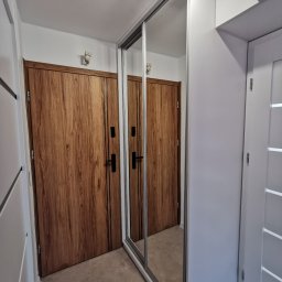 Najmart Sp. z o.o. - Nowoczesny przedpokój z przesuwną szafą wnękową z lustrem, odbijającą drewniane drzwi. Minimalistyczne oświetlenie punktowe i jasna kolorystyka ścian.