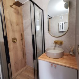 Najmart Sp. z o.o. - Nowoczesna łazienka z kabiną prysznicową typu walk-in, umywalką nablatową na drewnianym blacie i złotymi akcentami armatury.