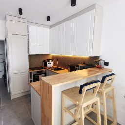 Najmart Sp. z o.o. - Jasna kuchnia na wymiar z drewnianym blatem i barkiem. Białe szafki z frezowanymi frontami, drewniane krzesła barowe z niebieskimi poduszkami. Oświetlenie LED pod szafkami.