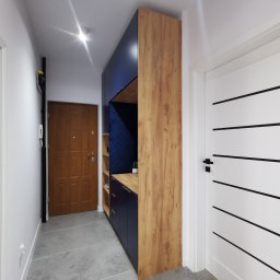 Najmart Sp. z o.o. - Nowoczesna zabudowa przedpokoju z drewnianym dekorem i granatowymi elementami, w tle białe drzwi z czarnymi akcentami i lustro. Minimalistyczny design.