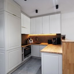 Najmart Sp. z o.o. - Jasna kuchnia z białymi szafkami i drewnianym blatem. Uwagę przykuwa złota armatura i oświetlenie LED pod szafkami, które podkreśla dekoracyjny panel ścienny.