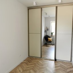 Najmart Sp. z o.o. - Jasna szafa przesuwna z lustrem w minimalistycznym wnętrzu. Parkiet w jodełkę, białe ściany i drzwi. W lustrze odbija się krzesło i roślina w doniczce.