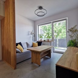 Jasne wnętrze z drewnianym ażurowym przepierzeniem, sofą z poduszkami, stolikiem kawowym i komodą. Za oknem zieleń. Nowoczesny design.