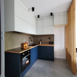 Nowoczesna kuchnia z białymi szafkami górnymi i granatowymi dolnymi, drewnianym blatem i geometrycznym wzorem na ścianie. Widoczny piekarnik i owoce w koszyku.