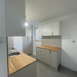 Jasna kuchnia z drewnianym blatem i złotymi uchwytami. Minimalistyczny design, szare płytki podłogowe. Widok na okno i szafę. Nowoczesne wykończenie.