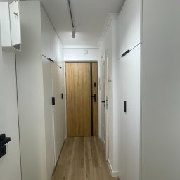 Białe szafy wnękowe z czarnymi uchwytami, zabudowa od podłogi do sufitu w wąskim korytarzu z drewnianą podłogą i drzwiami. Minimalistyczny design.