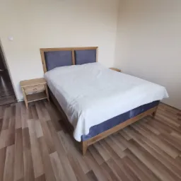 Sypialnia z drewnianym łóżkiem z szarym tapicerowanym wezgłowiem i szafką nocną, na podłodze panele imitujące drewno, widok na otwarte drzwi.