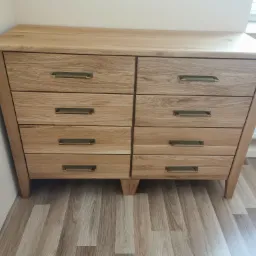 Drewniana komoda z sześcioma szufladami i prostokątnymi, metalowymi uchwytami, ustawiona na drewnianej podłodze przy białej ścianie.