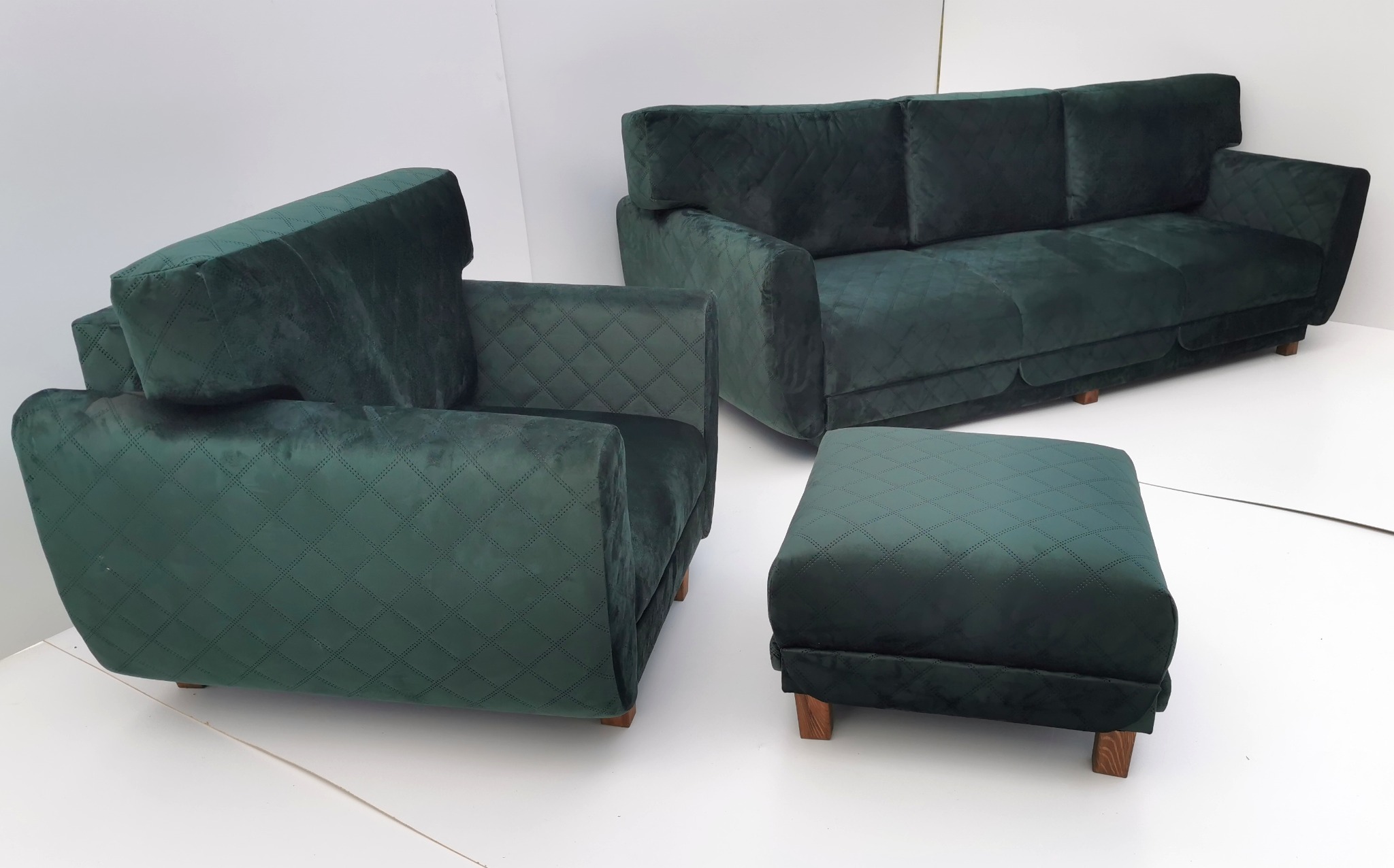 Komplet wypoczynkowy: sofa, fotel i pufa w odcieniach butelkowej zieleni, z pikowaniem w romby, na tle białej ściany. Minimalistyczny design, drewniane nóżki.