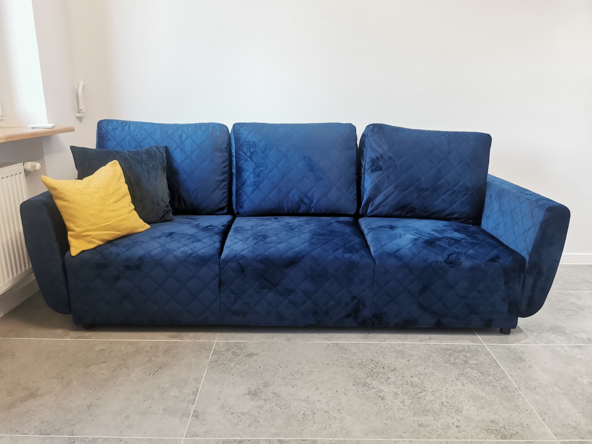 Granatowa sofa z pikowaniem w romby, z żółtą i granatową poduszką. Ustawiona na szarych płytkach, na tle białej ściany. Widoczny fragment grzejnika i parapetu.