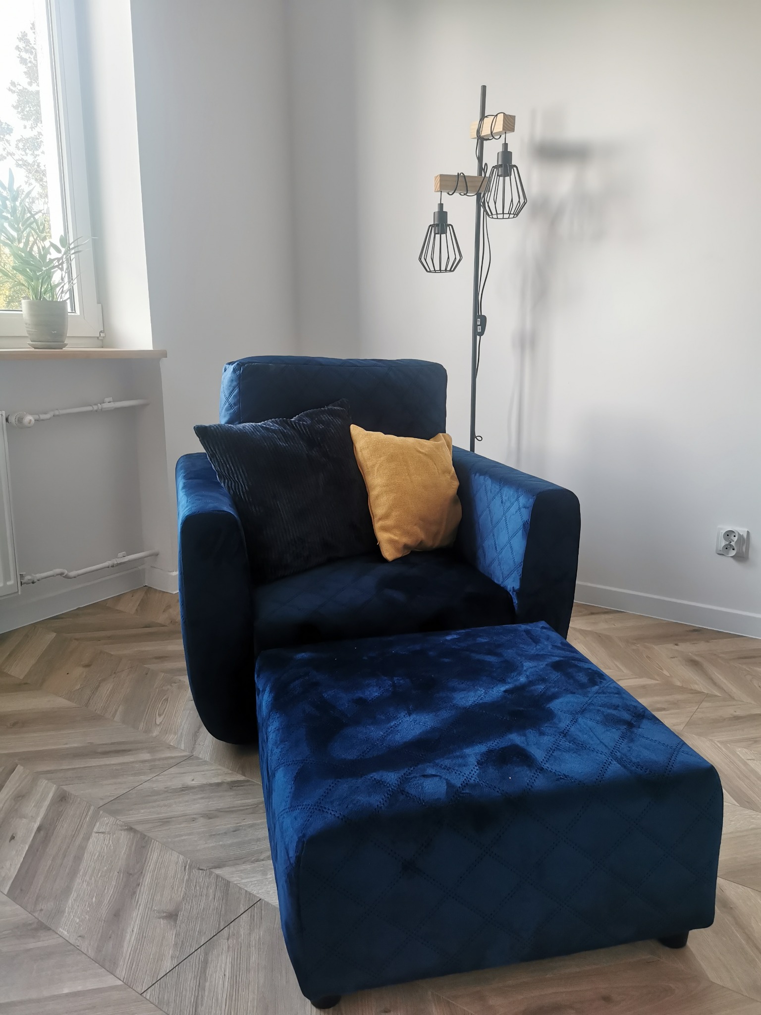 Granatowy fotel tapicerowany z podnóżkiem, zdobiony pikowaniem, z poduszkami w odcieniach granatu i złota, na parkiecie w jodełkę, oświetlony lampą w stylu industrialnym.