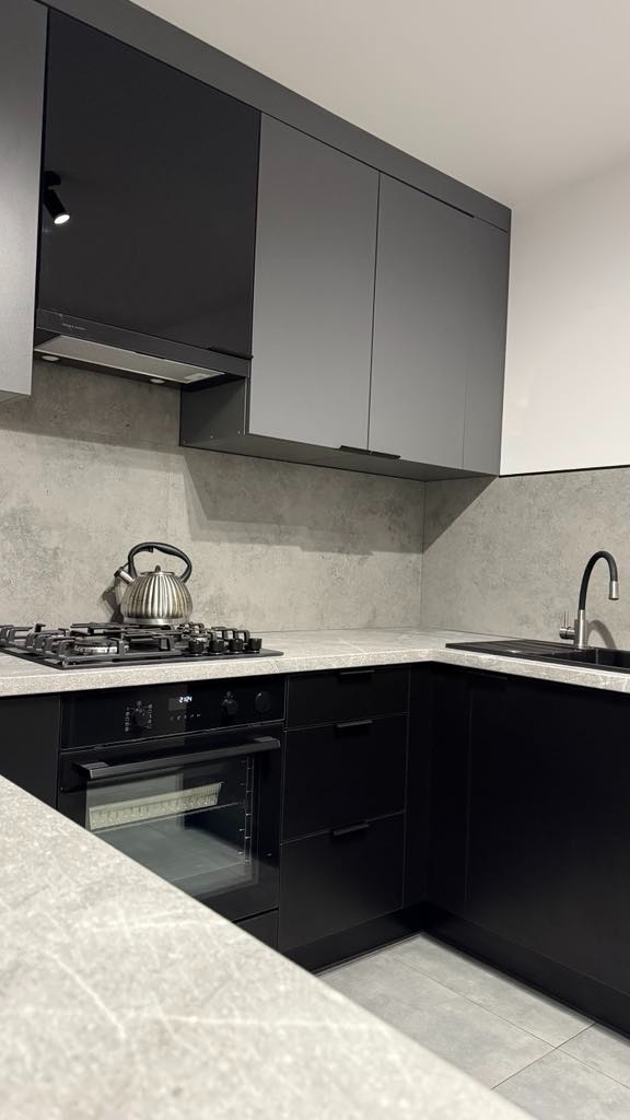 Nowoczesna kuchnia z szarymi i czarnymi szafkami, blatem imitującym kamień i srebrnym czajnikiem na płycie gazowej. Minimalistyczny design, oświetlenie punktowe.