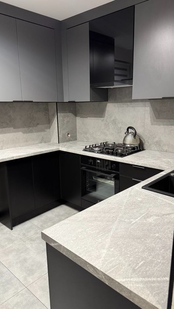 Nowoczesna kuchnia w odcieniach szarości i czerni z kamiennym blatem, minimalistycznymi szafkami i płytą grzewczą z czajnikiem. Stonowana kolorystyka, proste linie.