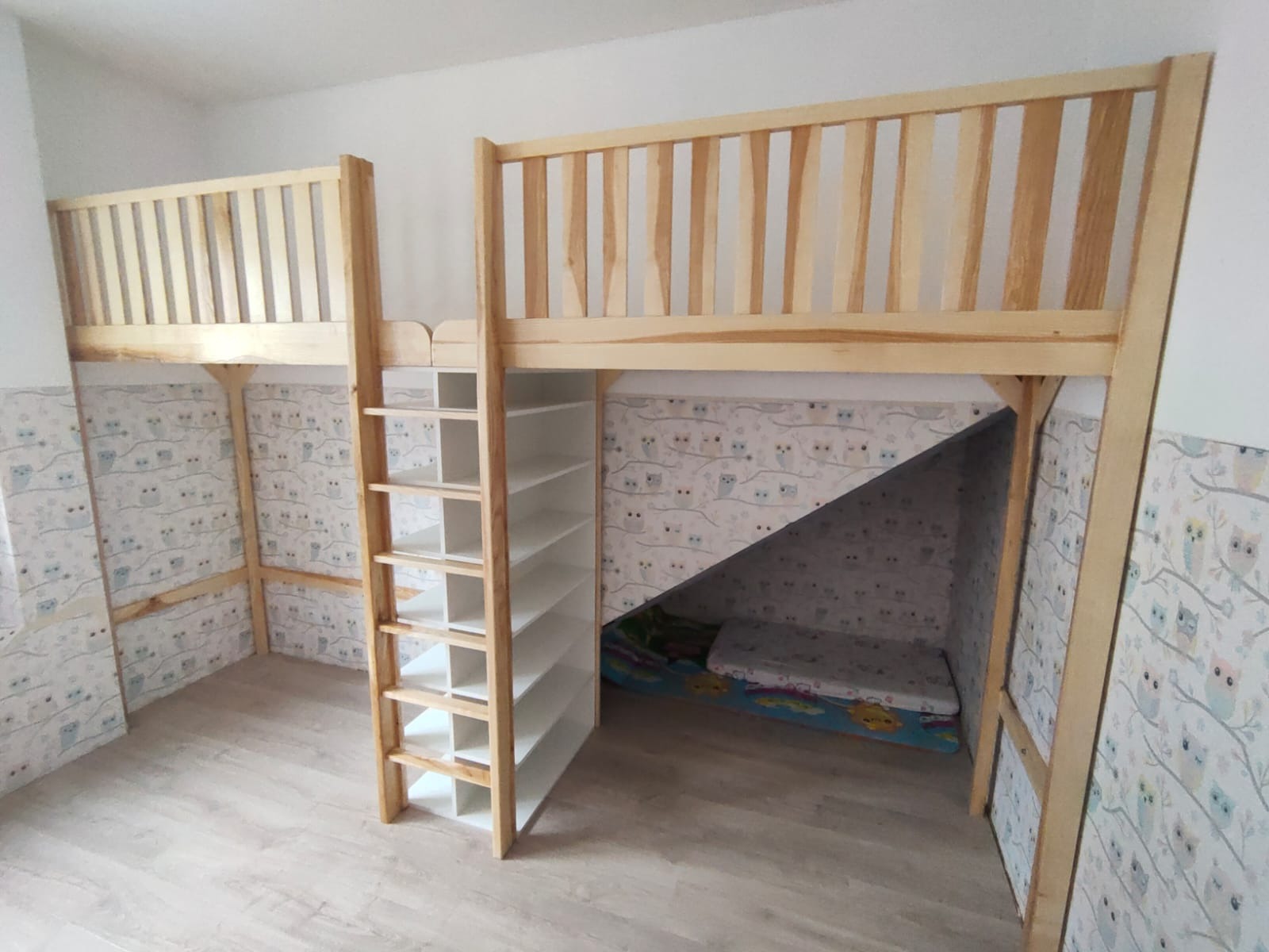 Drewniane łóżko na antresoli z drabinką i regałem, wykończone w jasnym drewnie, z przestrzenią do zabawy pod spodem, tapeta w sowy w tle.