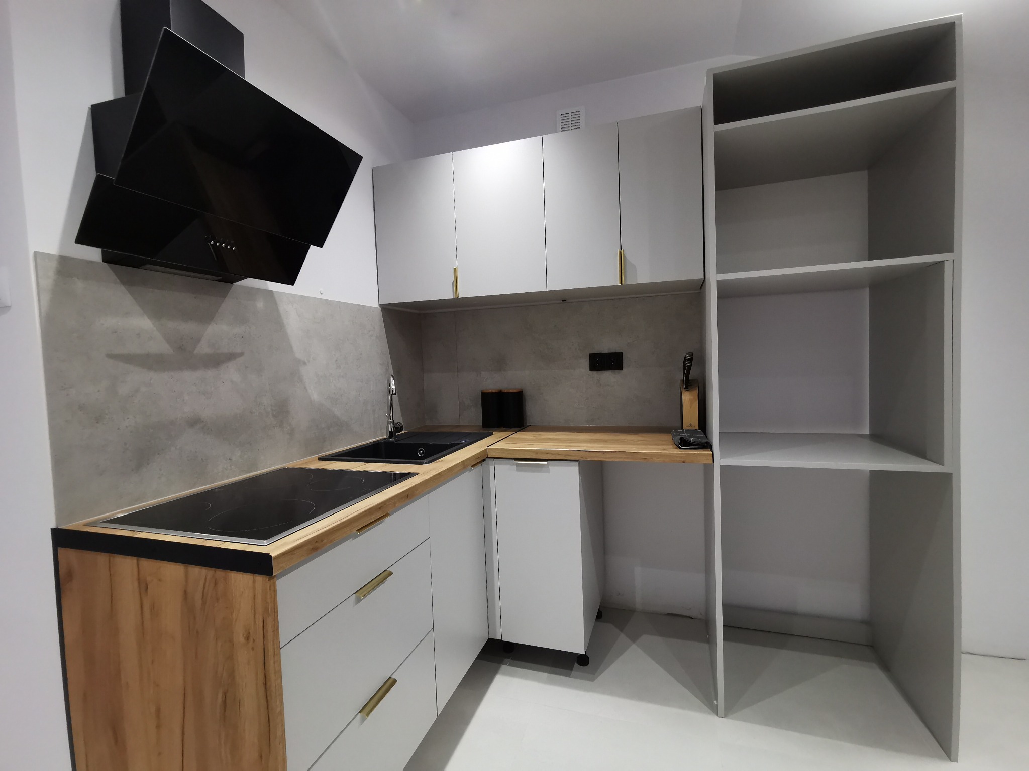 Nowoczesna kuchnia w kształcie litery L z białymi szafkami, drewnianym blatem i czarnym okapem. Minimalistyczny design z złotymi uchwytami i otwartą półką.