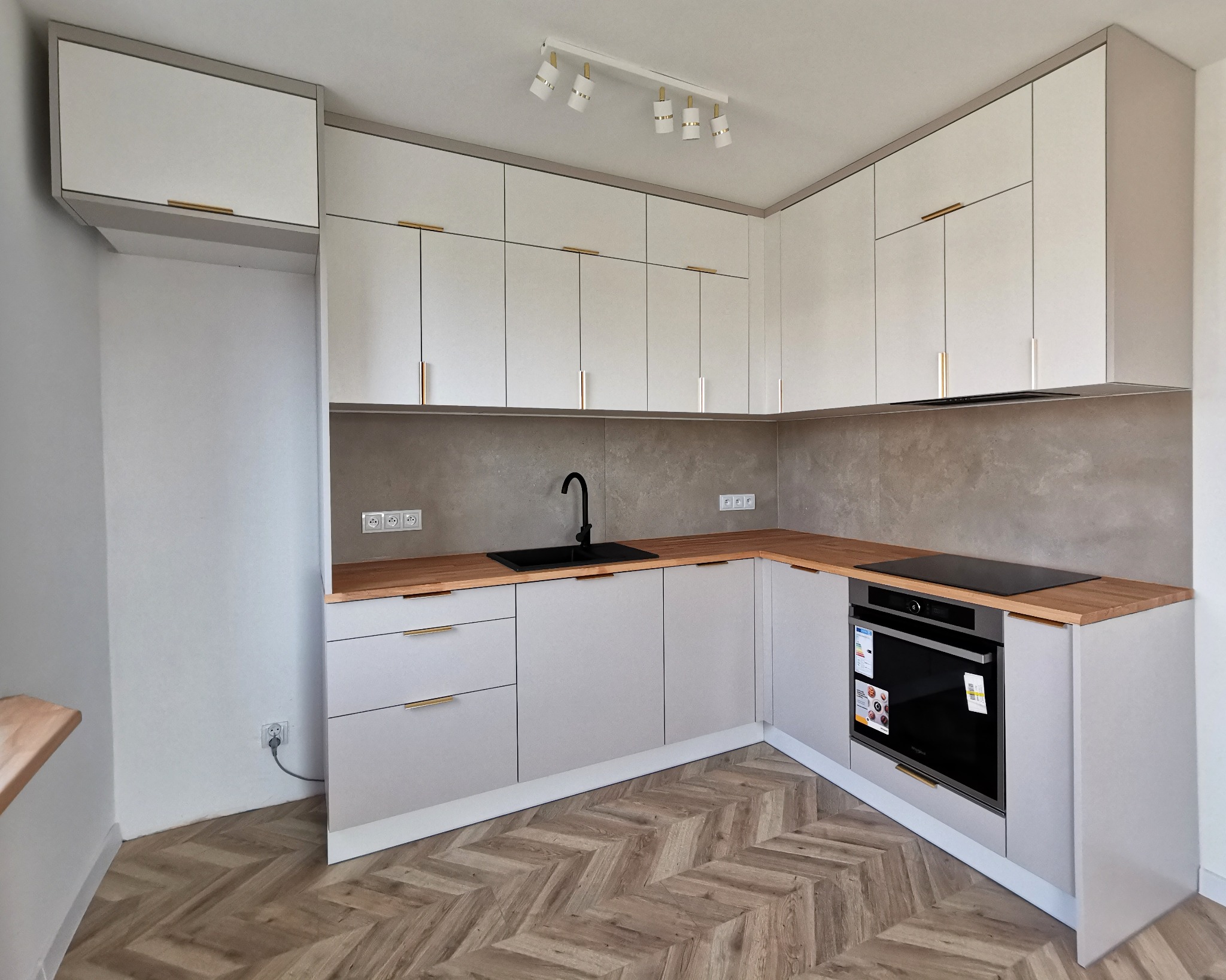 Jasna kuchnia z drewnianym blatem i złotymi uchwytami, oświetlona lampą sufitową. Na podłodze parkiet w jodełkę. Minimalistyczny design, nowoczesne wykończenie.