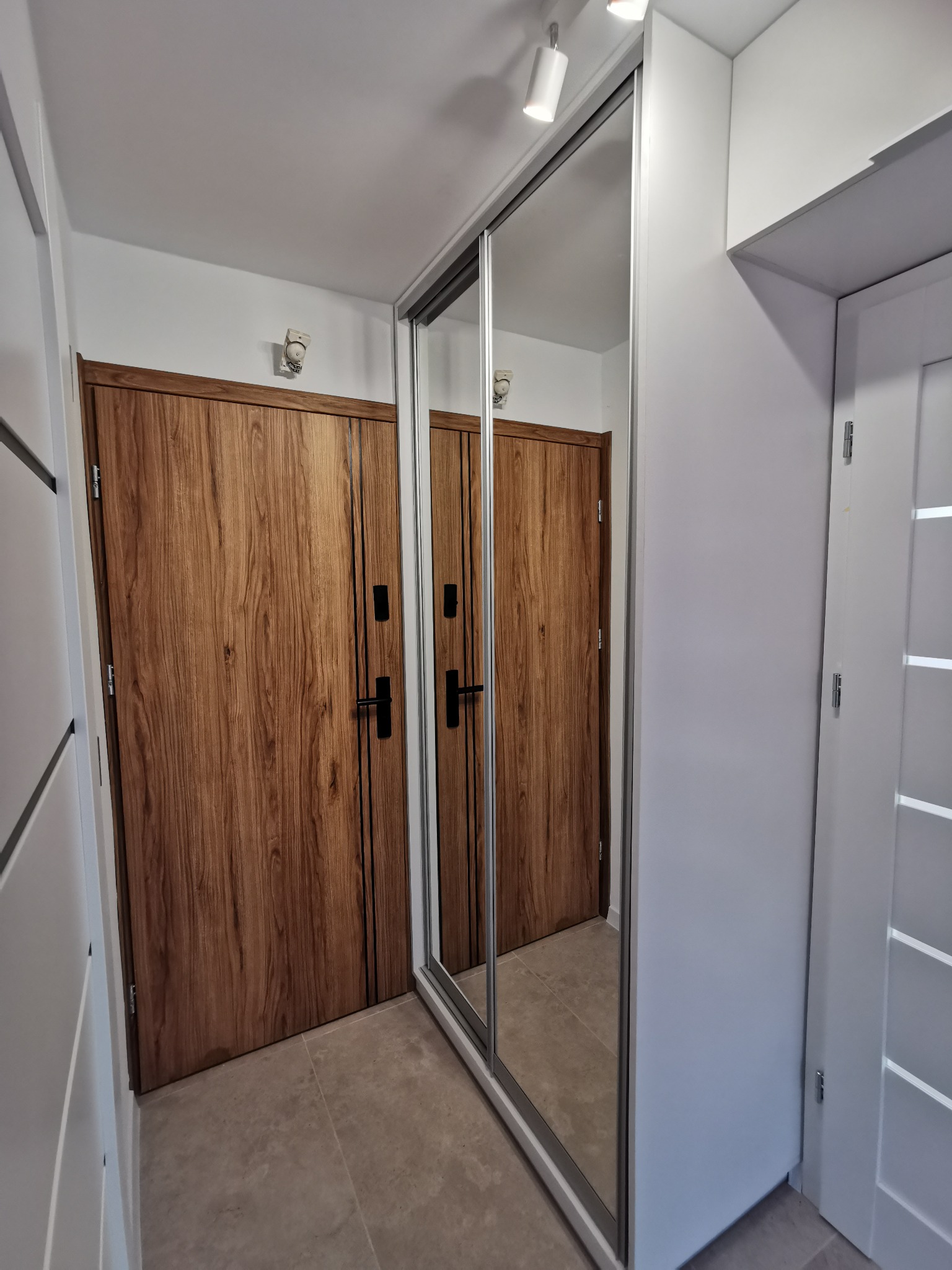 Nowoczesny przedpokój z przesuwną szafą wnękową z lustrem, odbijającą drewniane drzwi. Minimalistyczne oświetlenie punktowe i jasna kolorystyka ścian.