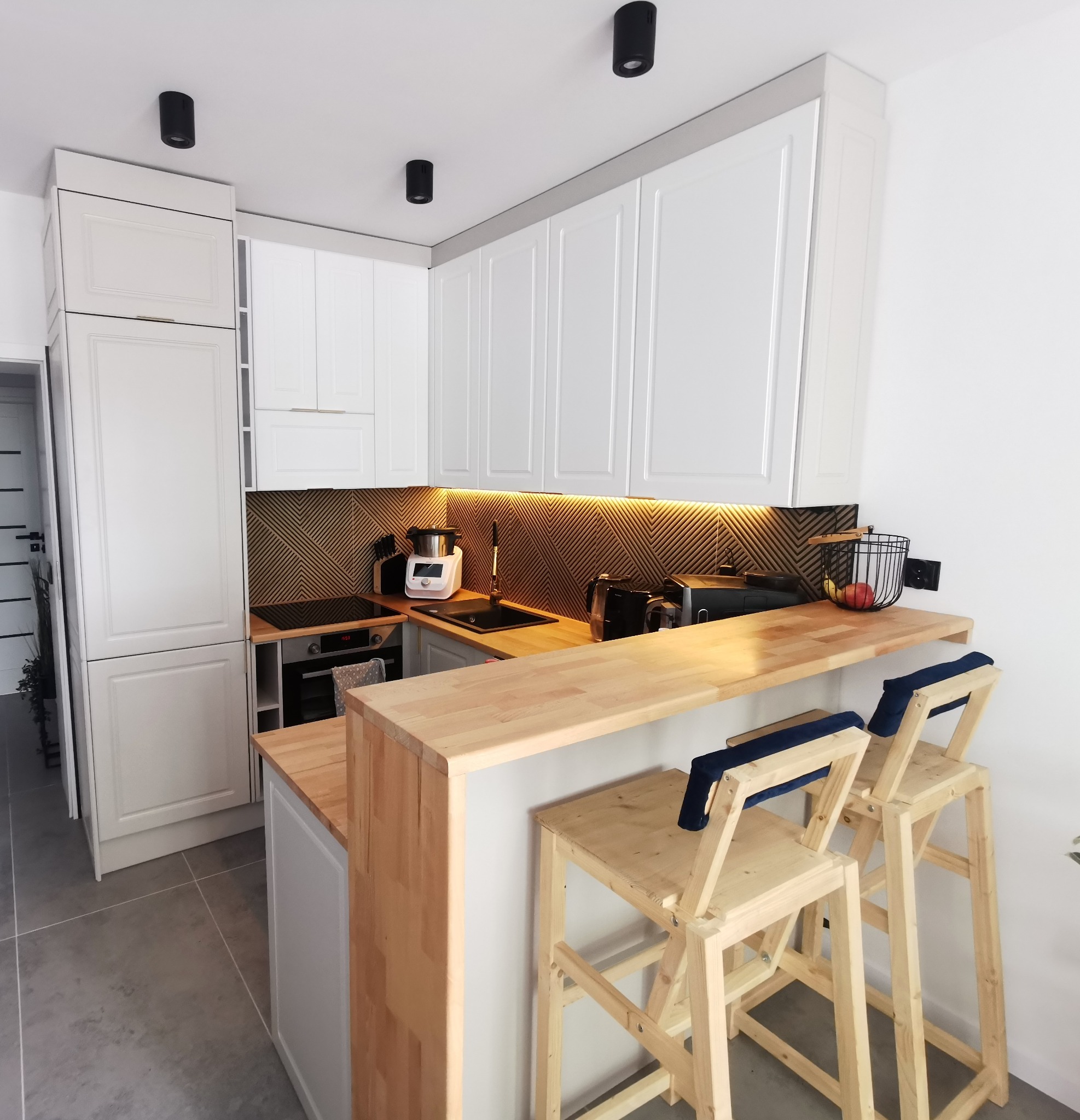 Jasna kuchnia na wymiar z drewnianym blatem i barkiem. Białe szafki z frezowanymi frontami, drewniane krzesła barowe z niebieskimi poduszkami. Oświetlenie LED pod szafkami.
