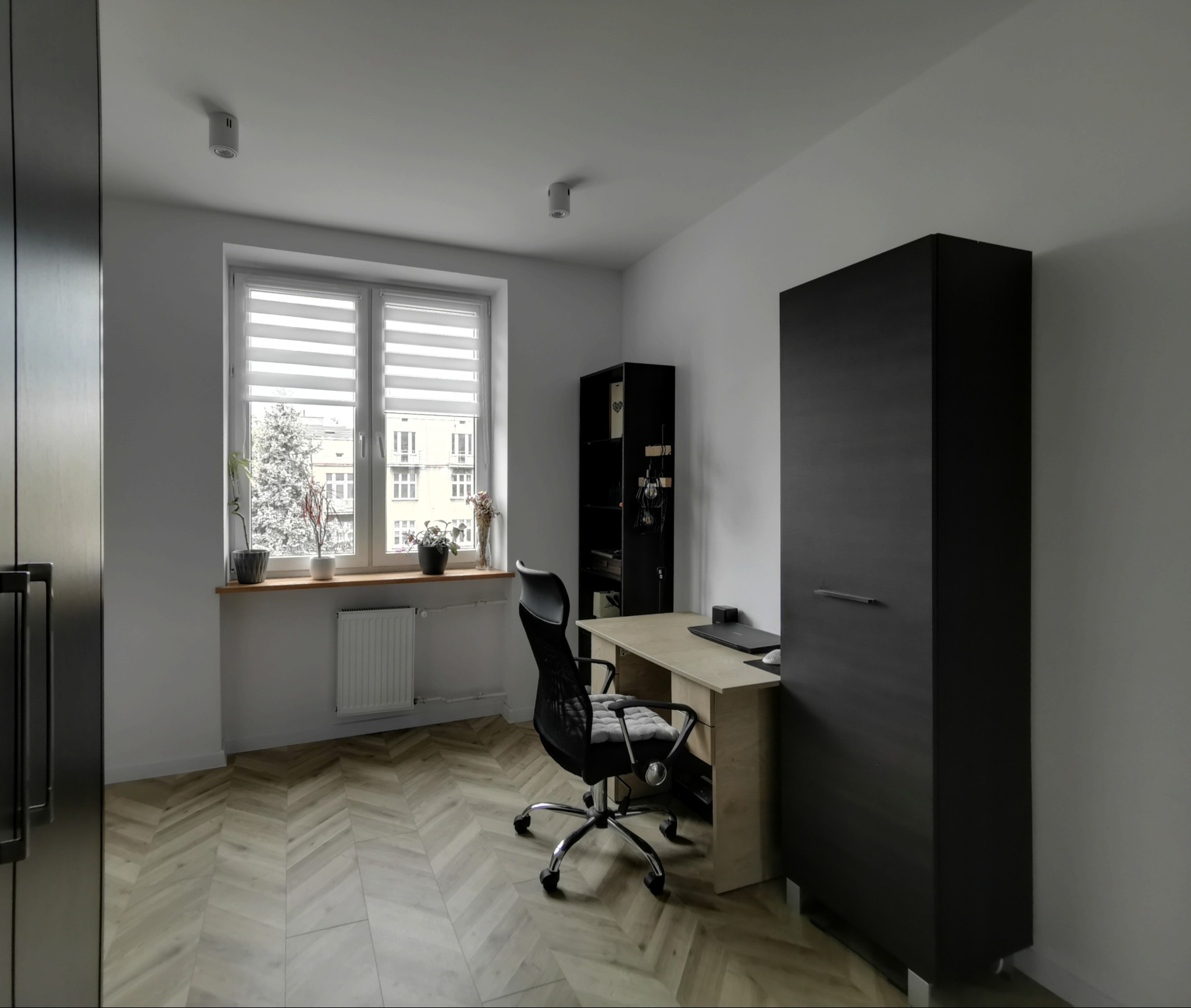 Nowoczesny pokój z biurkiem, krzesłem i szafą w ciemnym kolorze. Parkiet ułożony w jodełkę. Na parapecie rośliny doniczkowe. Minimalistyczne oświetlenie sufitowe.