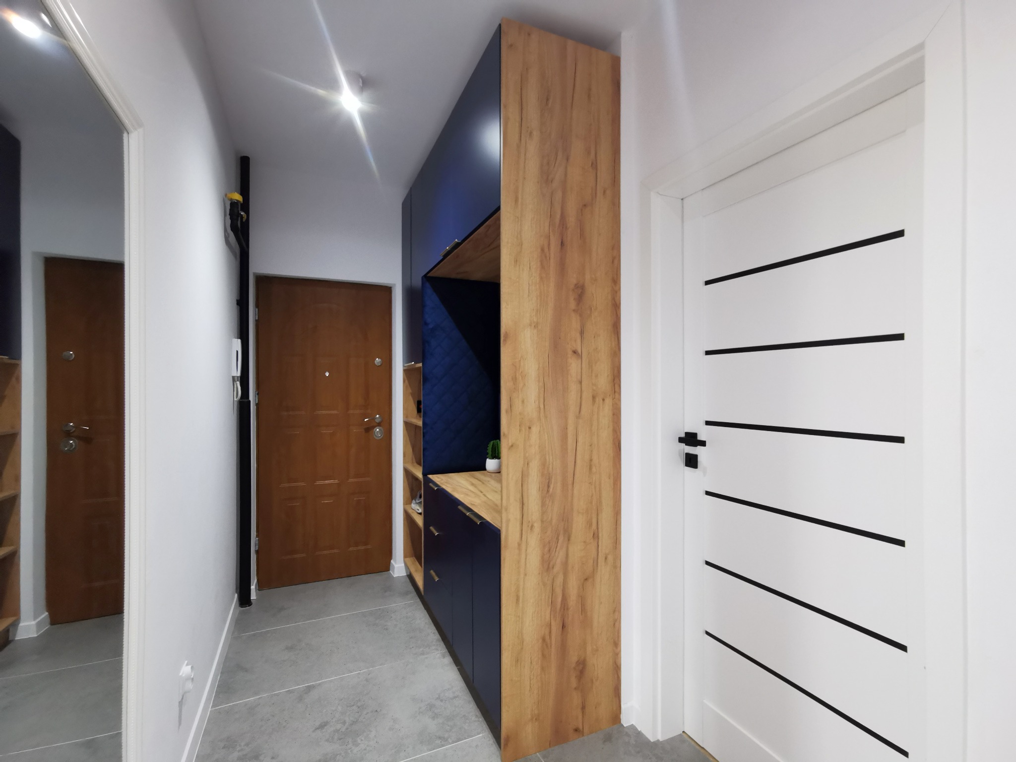 Nowoczesna zabudowa przedpokoju z drewnianym dekorem i granatowymi elementami, w tle białe drzwi z czarnymi akcentami i lustro. Minimalistyczny design.